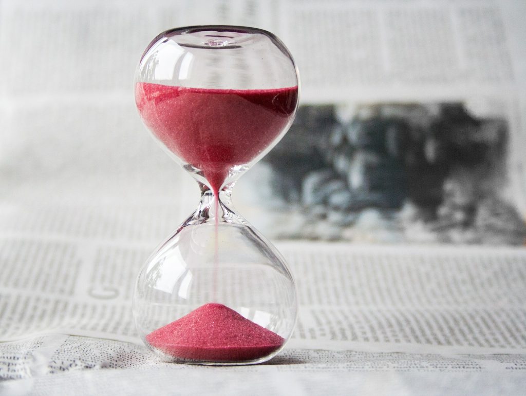 An hourglass filled with red sand, positioned on a surface with newspaper in the background. The sand is flowing from the upper to the lower bulb, indicating the passage of time. This image relates to the theme: 'How Long Does It Take To Learn Spanish.'