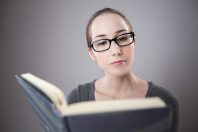 A young woman with glasses reading a book attentively against a gray background. The focus is on her expression as she engages with the content. This image relates to the concepts of 'Saber vs Conocer' in learning contexts.