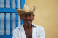 An older man wearing a straw cowboy hat and a white shirt, sitting against a yellow wall with blue window shutters. He has a cigar in his mouth, depicting traditional Cuban culture and lifestyle.