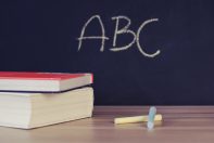 Two stacked books on a desk with colorful chalk pieces beside them. In the background, the letters 'ABC' are written on a black chalkboard, suggesting a learning environment focused on education.