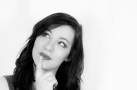 A woman with long dark hair, looking upward with a thoughtful expression while touching her finger to her chin. The image is in black and white.