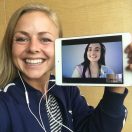 A woman smiling while holding a tablet that displays another person on a video call, both wearing headphones. The setting appears to be indoors with a wooden background, showing a lively atmosphere conducive to learning Spanish.