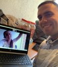 A man smiling while holding a laptop showing a video call with a woman who is waving. The scene includes a cluttered table with items like jars and a jacket in the background. This image could support a theme on how to learn Spanish through video chats.