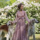 A woman in a light pink flowing dress standing in a garden with two Borzoi dogs. The background features blooming flowers and greenery, highlighting the serene outdoor setting.