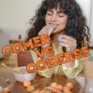 A woman with curly hair smiling while eating from a bowl of fruit on a wooden table, with the text 'COMER VS COMERSE' displayed above her in a bold font. The setting is bright and modern, with soft furnishings in the background.