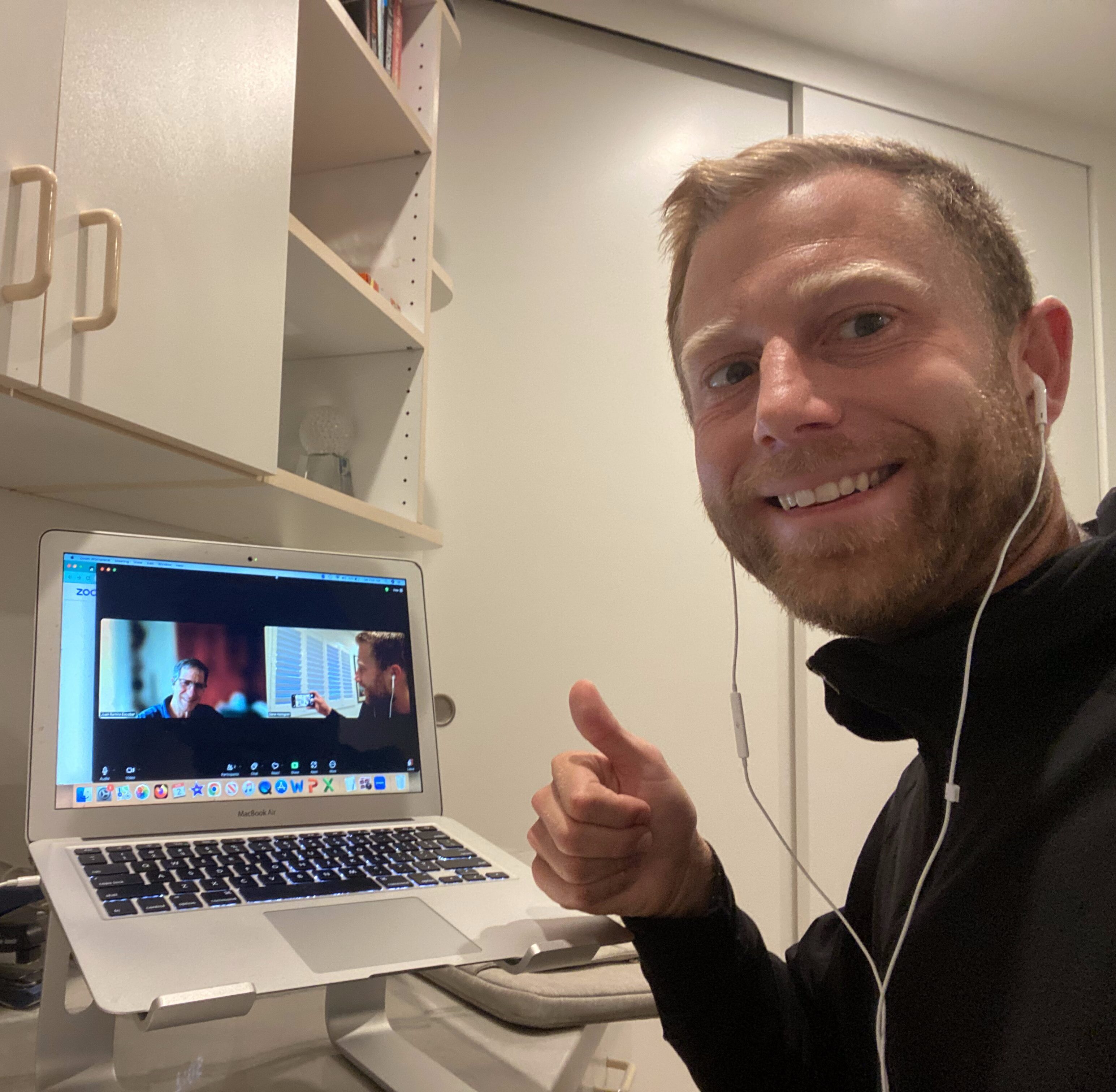 A man with short hair and a beard, wearing a black shirt, gives a thumbs up while sitting at a desk with an open laptop displaying a screen. The background includes shelves and cabinets.