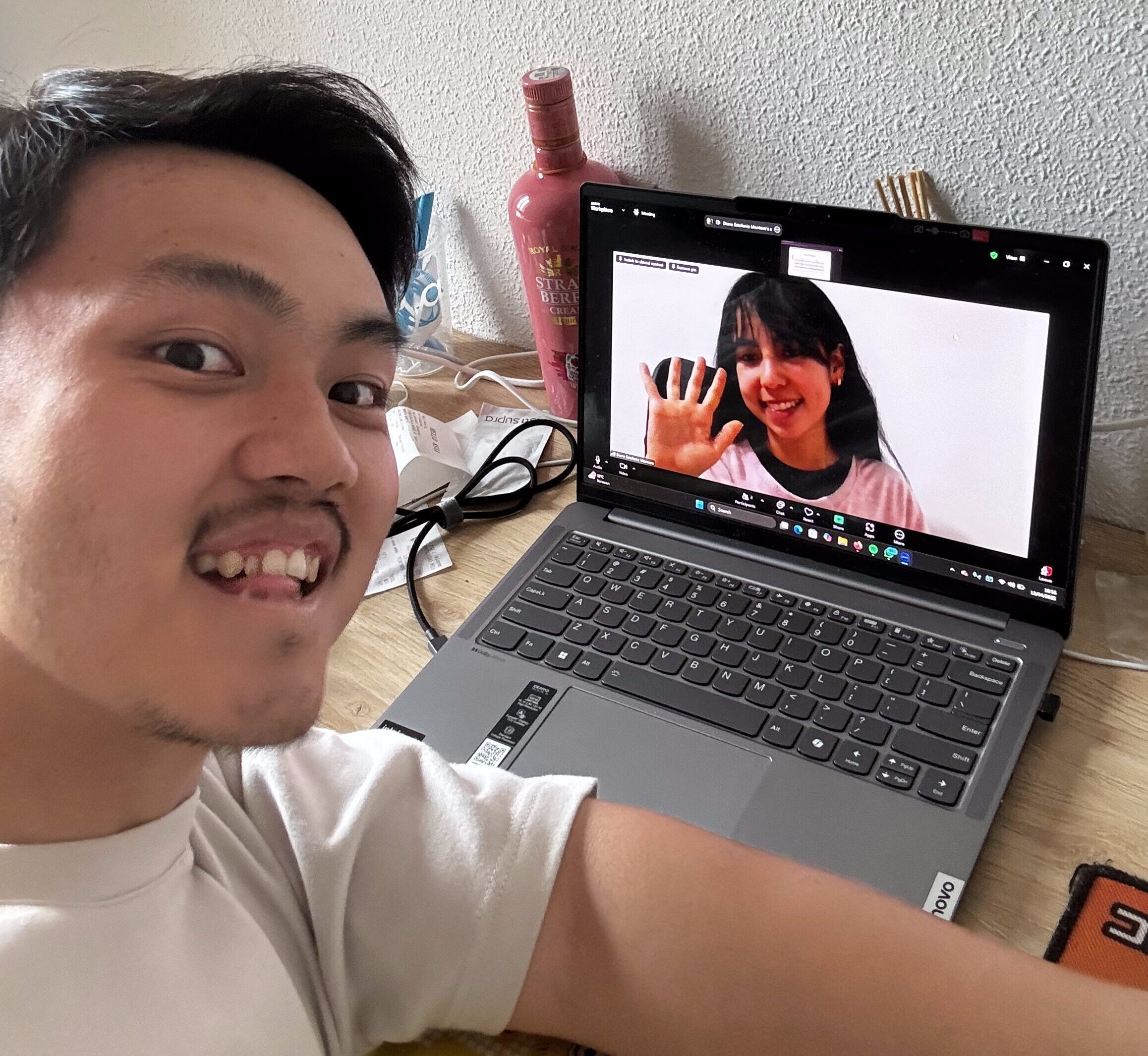A young man smiling and taking a selfie while attending a virtual meeting on his laptop. The screen shows a woman waving, suggesting a friendly interaction. The workspace includes wires and a bottle in the background.