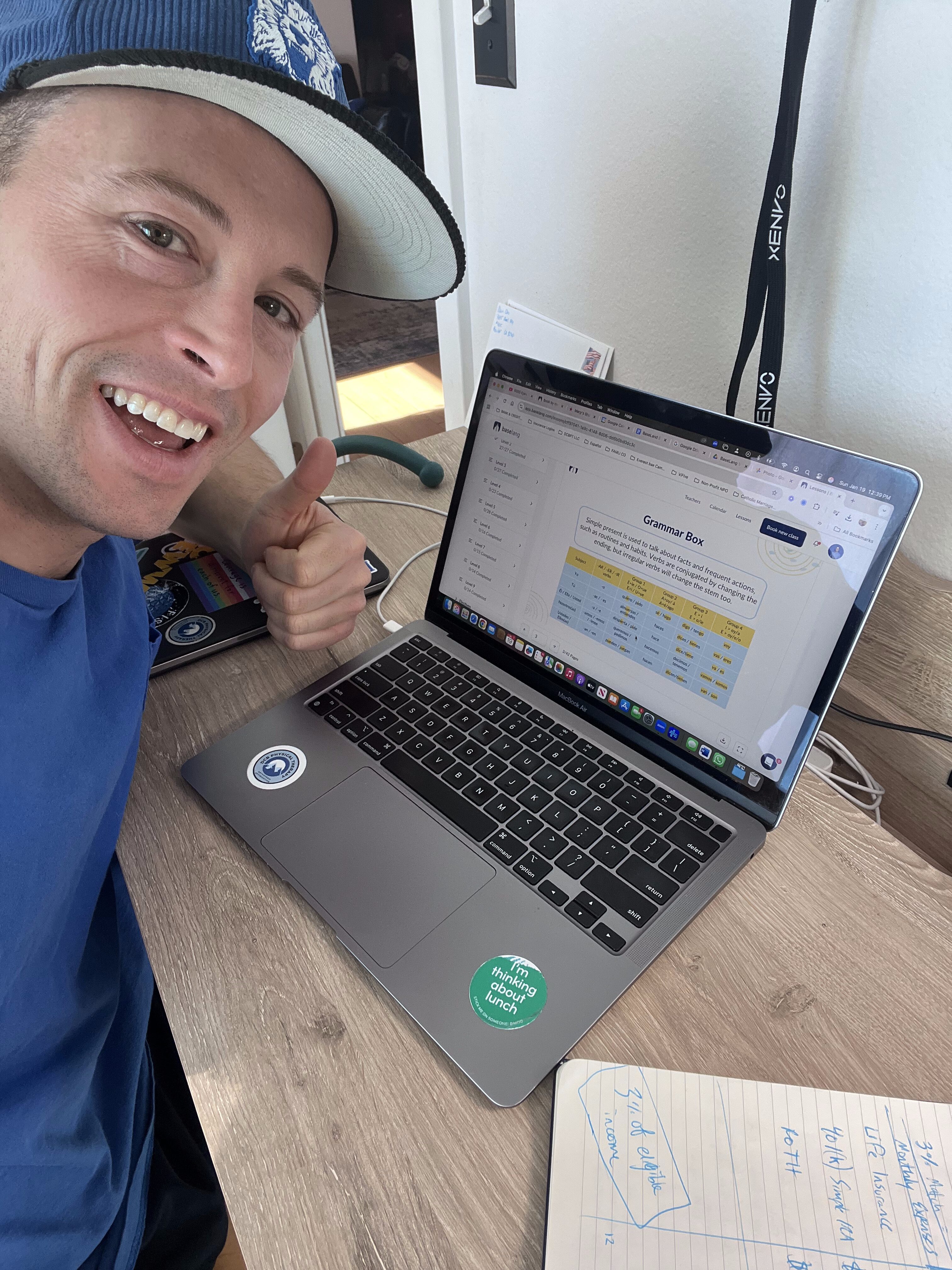 A person smiling and giving a thumbs up while sitting at a desk with a laptop displaying a spreadsheet. There is a notebook and writing materials nearby, indicating engagement in learning Spanish.