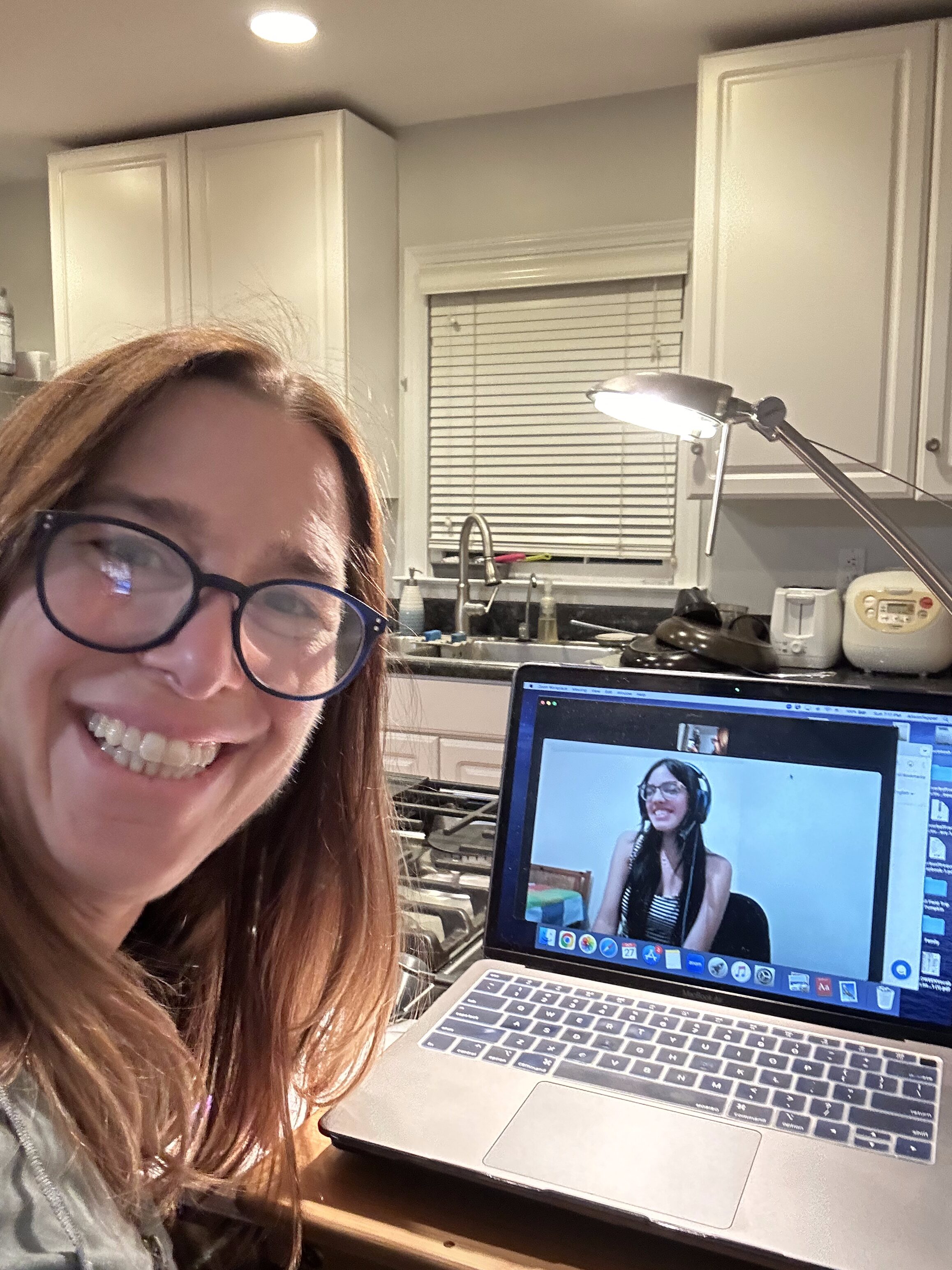 A woman smiling at the camera while sitting at a kitchen table, with a laptop open in front of her displaying a video call with another person. The kitchen has white cabinets and various utensils in the background.