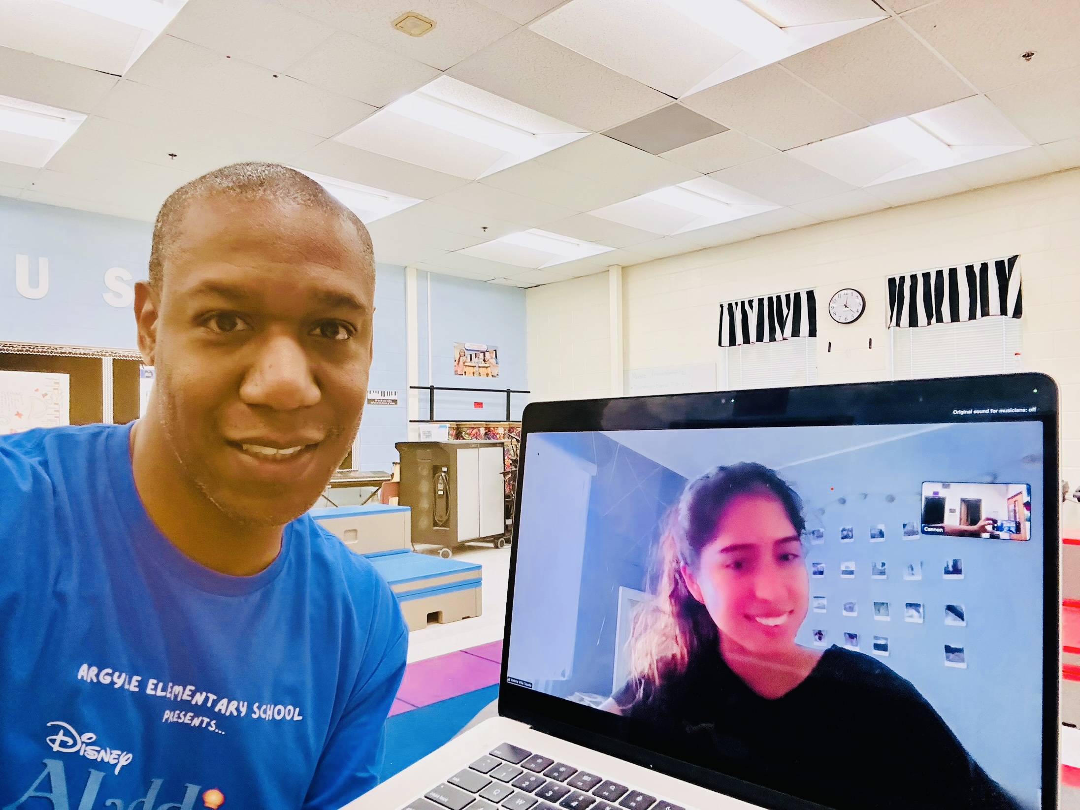 A man in a blue shirt sits at a table with a laptop open, displaying a video call with a woman smiling. The background shows a bright, spacious room with gym mats and a clock on the wall.
