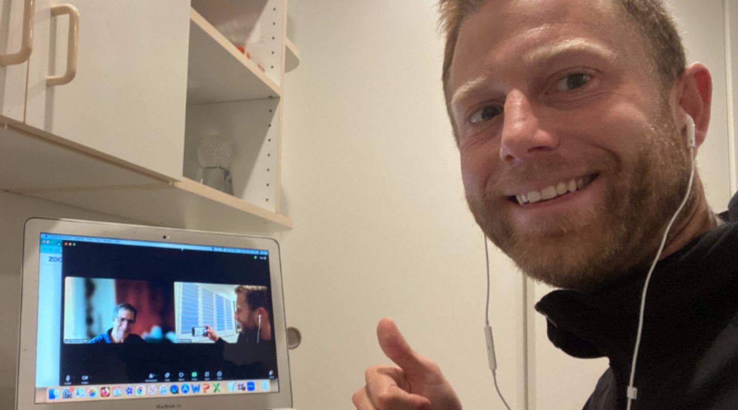 A man smiling and giving a thumbs up while participating in a video call on his laptop. The laptop screen displays another person in a video call, and the background features a simple, organized workspace.