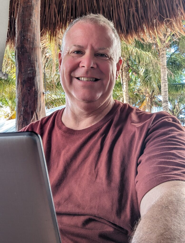 A man wearing a maroon shirt sits outside under a thatched roof, smiling at the camera while working on a laptop. Palm trees are visible in the background.