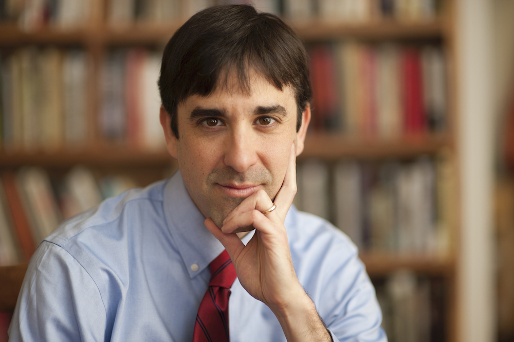 A man in a blue dress shirt and red tie sits with his chin resting on his hand, appearing thoughtful. Behind him is a blurred bookshelf filled with books.