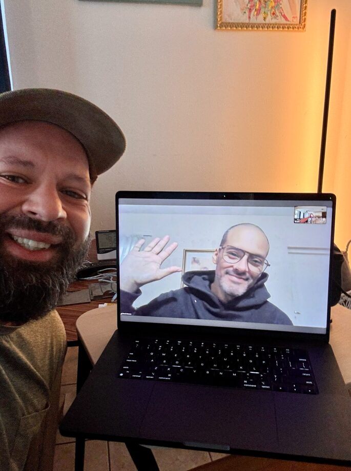 A smiling man with a beard taking a selfie while sitting in front of a laptop displaying another man who is waving. The scene is indoors with soft lighting.