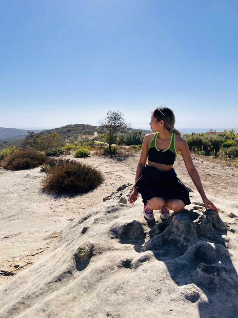 A woman in a black skirt and green top sitting on a rock, looking away. She is on a scenic hillside with sparse vegetation, under a clear blue sky.