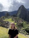 A person with long hair stands smiling in front of the ancient ruins of Machu Picchu, surrounded by lush greenery and steep mountains under a partly cloudy sky. The terraced structures of the ruins are visible in the background.