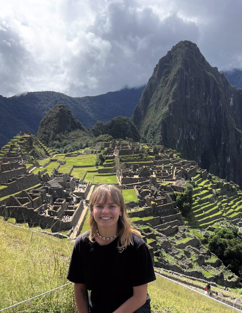A person with long hair stands smiling in front of the ancient ruins of Machu Picchu, surrounded by lush greenery and steep mountains under a partly cloudy sky. The terraced structures of the ruins are visible in the background.