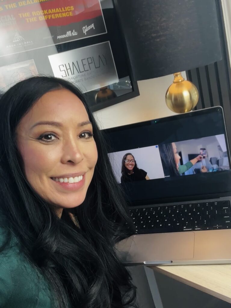 A woman with long black hair smiles at the camera while sitting at a desk with a laptop displaying a video call. The background features a lamp and framed posters on the wall.